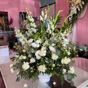 Large white and green floral arrangement composed of carnations, chrysanthemums, roses, and greenery in a white pedestal vase on a marble table with a pink wall and decor in the background.