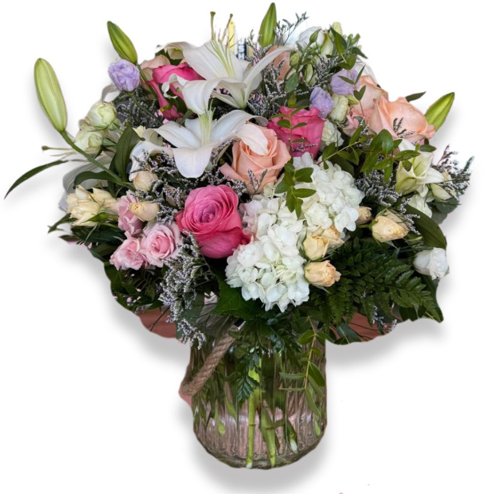 Blushing Elegance Bouquet featuring pink roses, white lilies, hydrangeas, and mixed seasonal flowers arranged in a clear glass vase with greenery, captured on a white background.