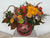 Festive Halloween-themed floral arrangement featuring a carved pumpkin jack-o'-lantern vase filled with orange, yellow, and purple flowers on a white background.